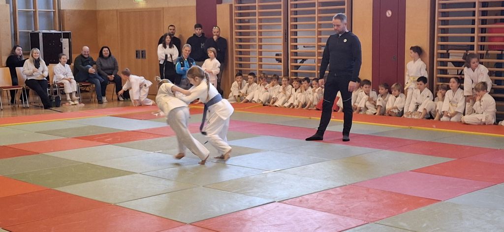 Judo-Kinder kämpfen auf einer Matte, während Zuschauer und Trainer zusehen.